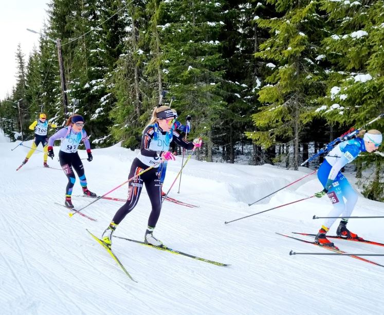 Bild på flera skidåkare i spåret med skog i bakgrunden. Biathlon Östersunds Nicolina Lindqvist i förgrunden i svart och orange tävlingsdräkt